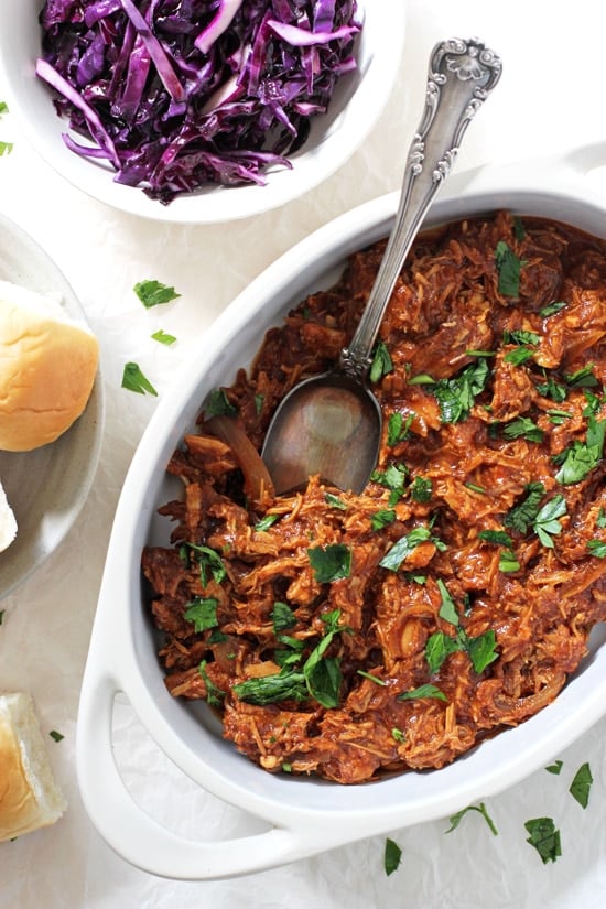 A white baking dish filled with Pulled BBQ Chicken with slaw and buns to the side.