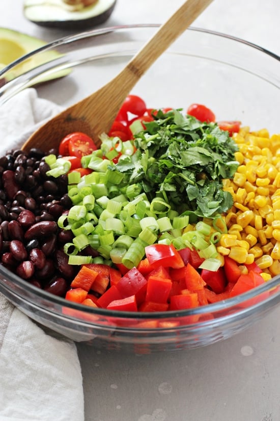 A glass bowl filled with all the components for Southwestern Bean Salad with a wooden spoon in the dish.