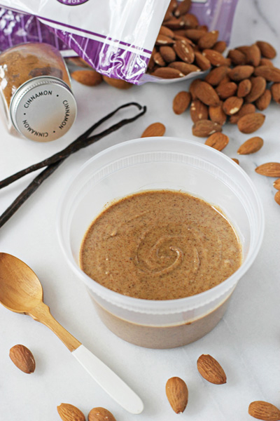 A container filled with Cinnamon Almond Butter with raw almonds scattered around.