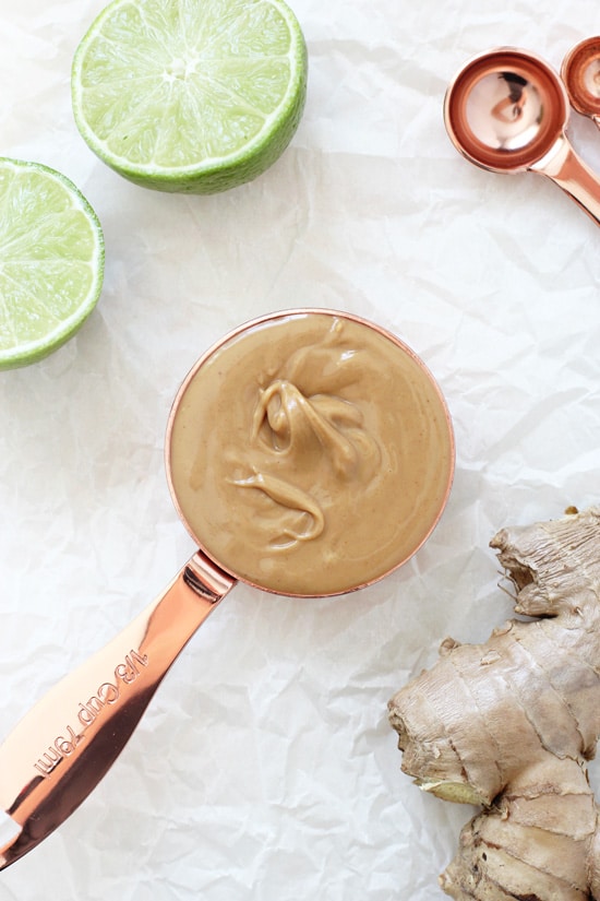 A measuring cup filled with peanut butter and fresh ginger and limes to the side.