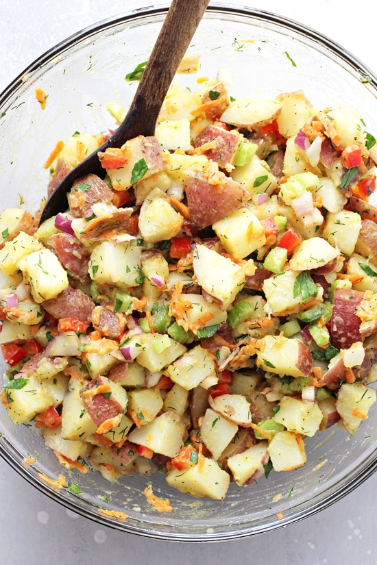 A glass mixing bowl filled with Healthy Potato Salad with a wooden spoon in the dish.