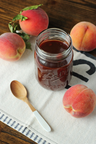 A jar of Peach Chipotle BBQ Sauce with fresh peaches scattered around.