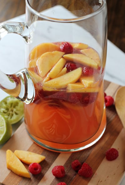 A glass pitcher of Peach and Raspberry Sangria on a wood cutting board.