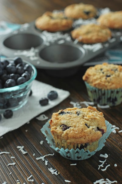 Two Coconut Blueberry Muffins on a wood surface with more to the side.