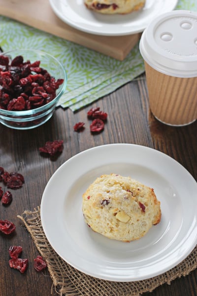 A Cranberry Orange Scone on a plate with coffee and dried cranberries to the side.