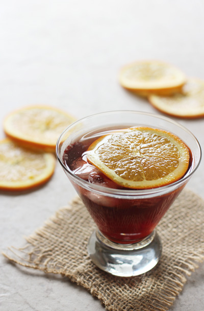A glass filled with Bourbon Pomegranate Cocktail with orange slices in the background.