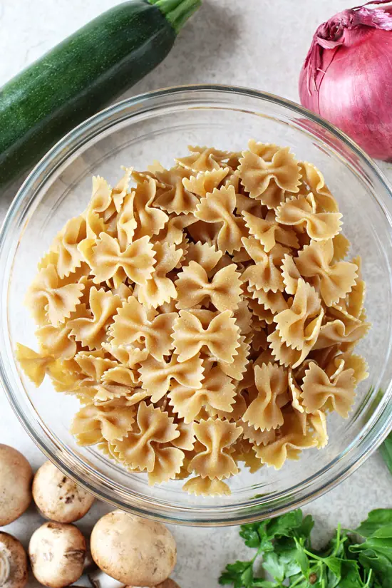 Dry pasta in a glass bowl with fresh veggies scattered around.