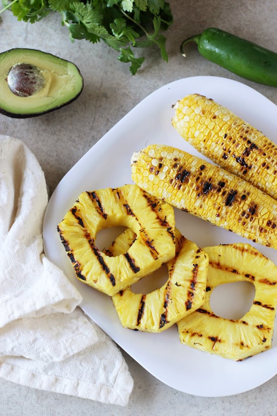 Grilled ears of corn and pineapple slices on a white plate.
