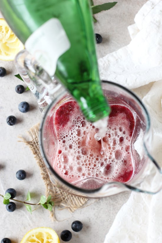 A pitcher filled with Blueberry Lemonade with someone pouring sparkling water in.