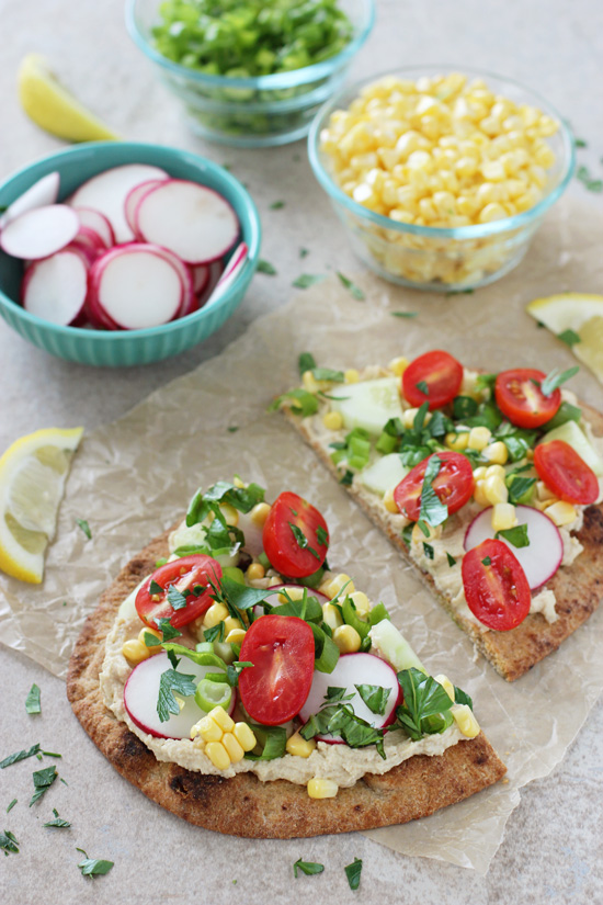 A Hummus Flatbread sliced in half with chopped veggies in bowls in the background.