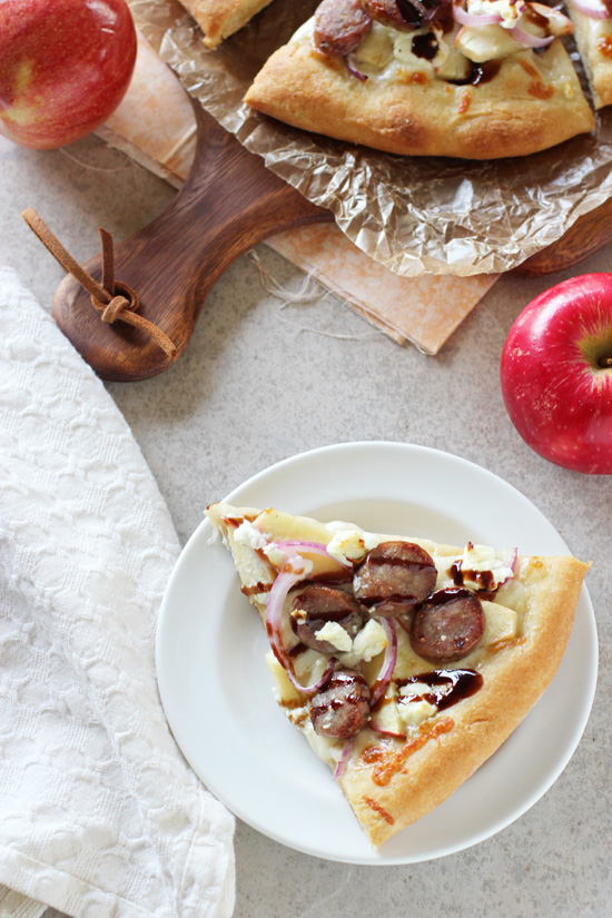 A slice of Bratwurst Pizza on a white plate with the full pie in the background.