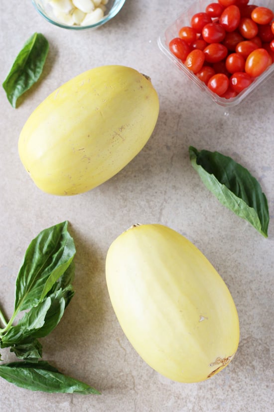 Two whole spaghetti squash, a pint of cherry tomatoes and fresh basil on a beige surface.