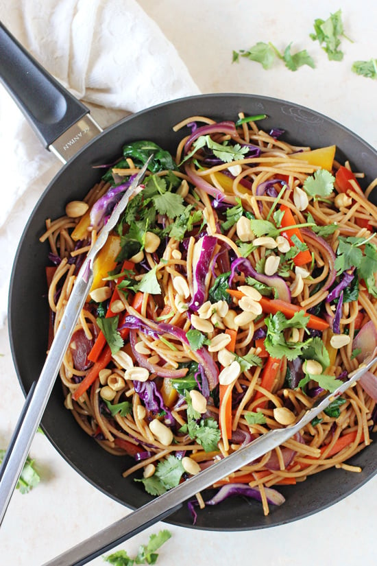A black skillet filled with Rainbow Vegetable Lo Mein with tongs in the pan.