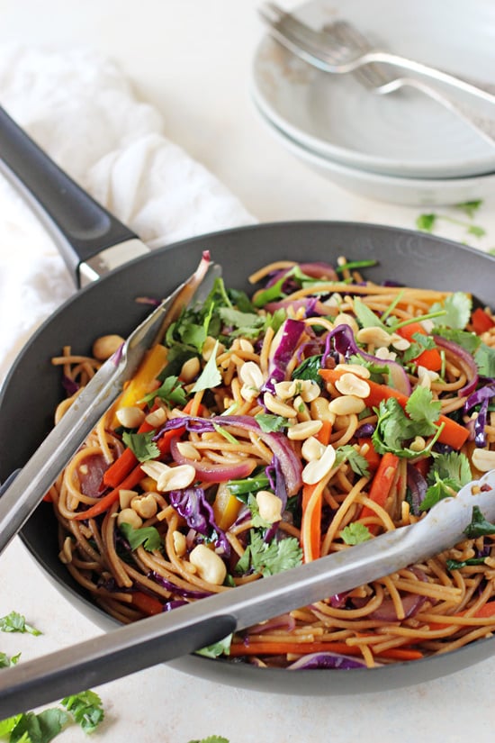 A black skillet filled with Veggie Lo Mein with tongs in the pan.