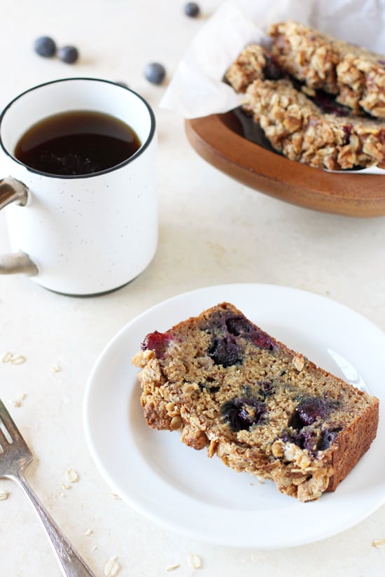 A slice of Blueberry Banana Bread on a white plate with coffee and more bread in the background.