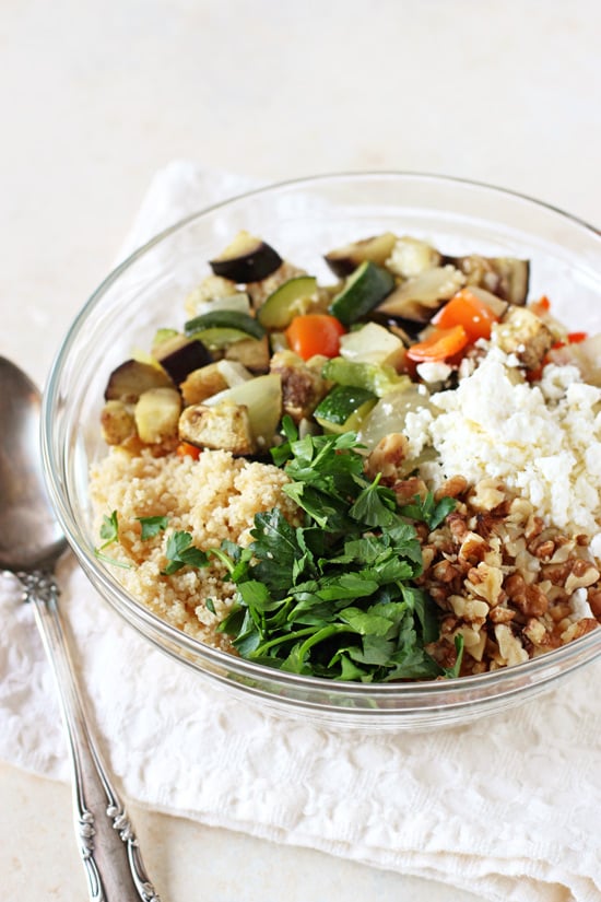 A glass bowl filled with couscous, roasted veggies, feta, walnuts and herbs.