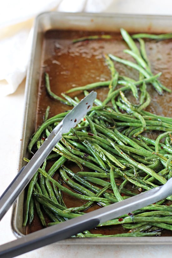 A baking sheet with Sweet and Spicy Green Beans and tongs.