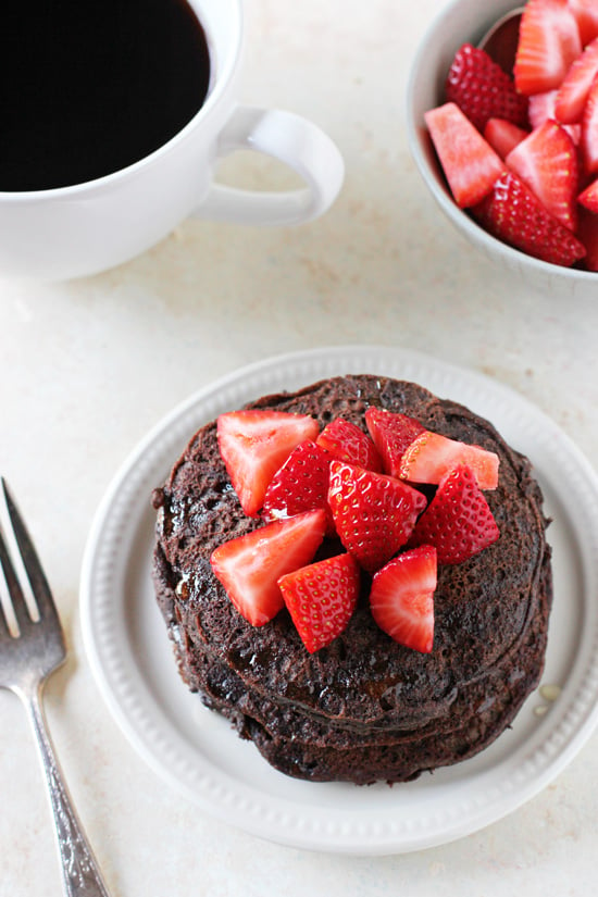 A stack of Chocolate Buckwheat Pancakes on a white plate with coffee and strawberries in the background.