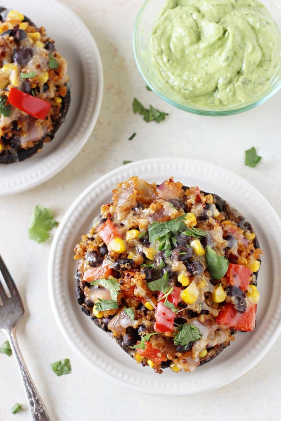 Two Mexican Quinoa Stuffed Portabella Mushrooms on white plates with a bowl of avocado sauce to the side.