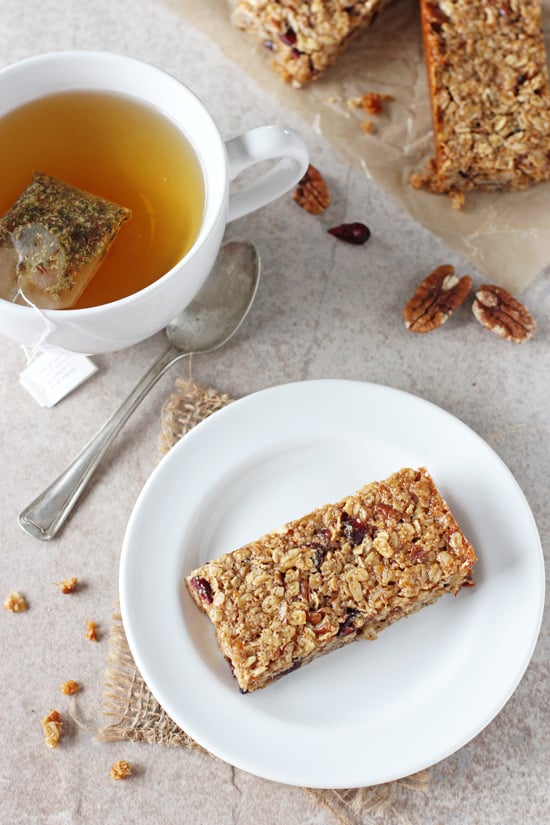 A white plate with a Cranberry Orange Granola Bar with tea and more granola bars in the background.