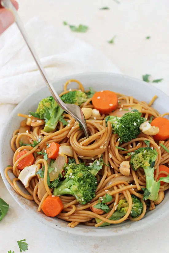 A grey bowl filled with Broccoli Carrot Noodle Stir-Fry with a hand twirling some around a fork.