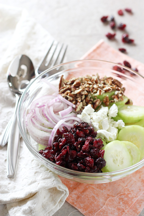 A glass bowl filled with Cranberry Pecan Cucumber Salad with a serving spoon and fork to the side.