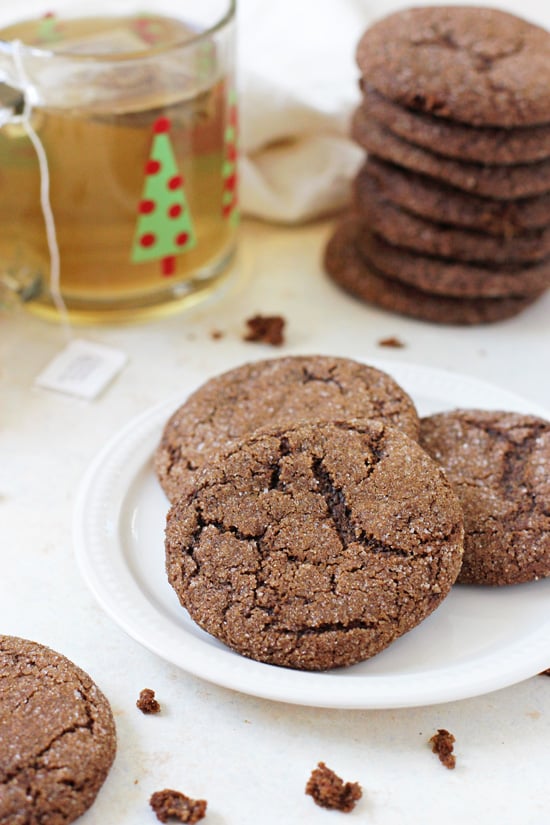 Three Soft & Chewy Whole Wheat Gingerbread Cookies on a white plate with more cookies and a cup of tea in the background.