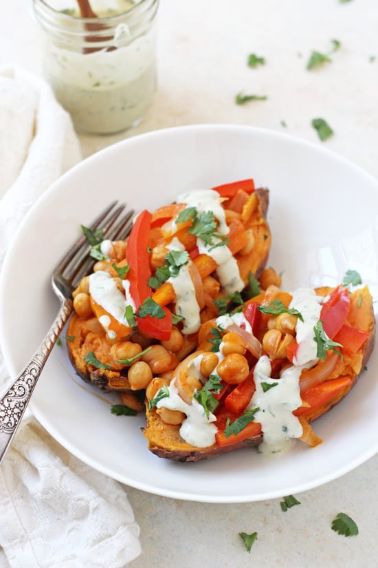 Side angle view of Buffalo Chickpea Stuffed Sweet Potatoes in a white bowl with a fork and a jar of tahini ranch in the background.