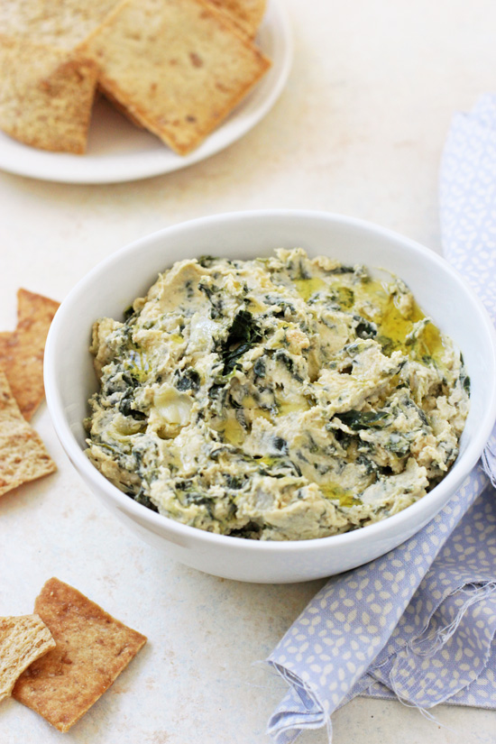 A bowl of Spinach Artichoke Hummus with pita chips and a blue napkin to the side.