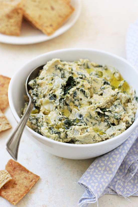 A white bowl filled with Spinach Artichoke Hummus with a silver spoon in the dish.