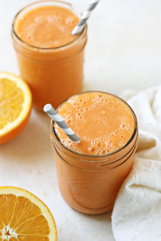 Two glasses filled with Orange Carrot Smoothie with grey straws and orange halves in the background.