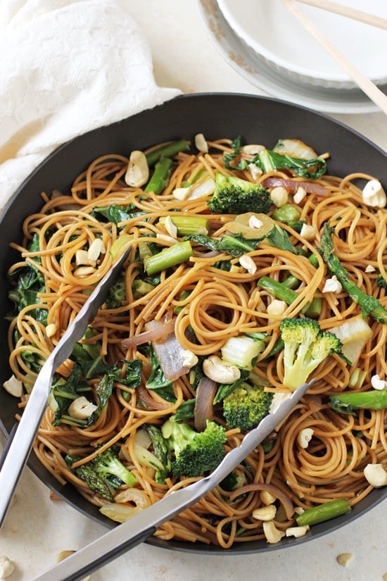 Vegan Lo Mein in a black skillet with tongs and plates in the background.