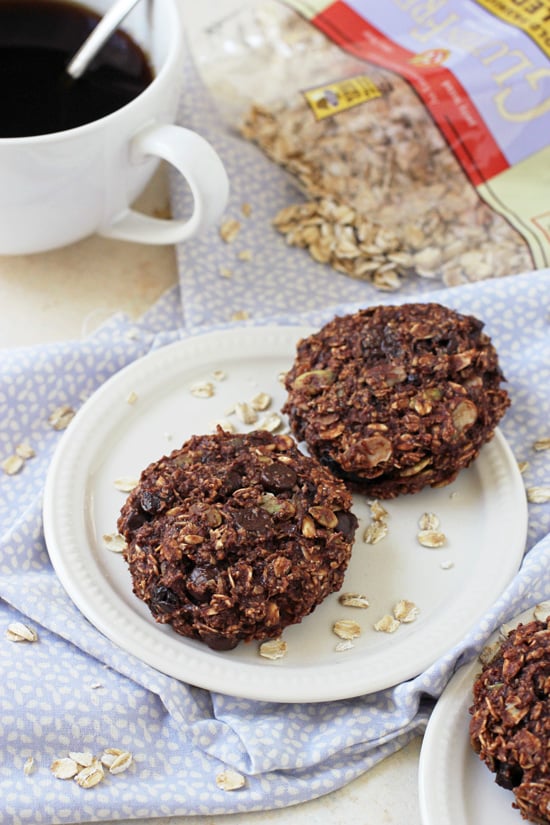 Side angle view of two Chocolate Breakfast Cookies on a plate with coffee and a bag of oats in the background.