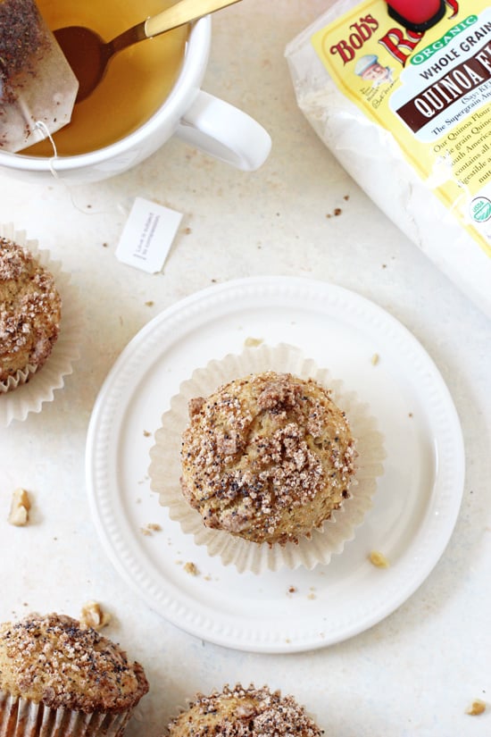 A Banana Bread Quinoa Muffin on a plate with a cup of tea and more muffins scattered around.