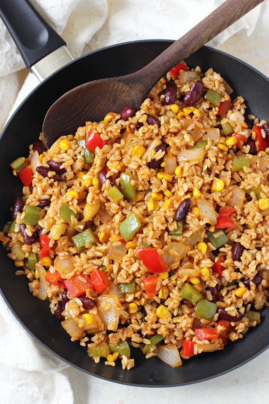 A skillet filled with Vegetable Cajun Brown Rice with a wooden spoon in the dish.