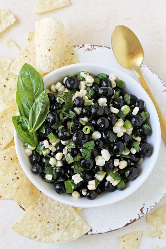 A white bowl filled with Blueberry Corn Salsa with tortilla chips and a gold spoon on the side.