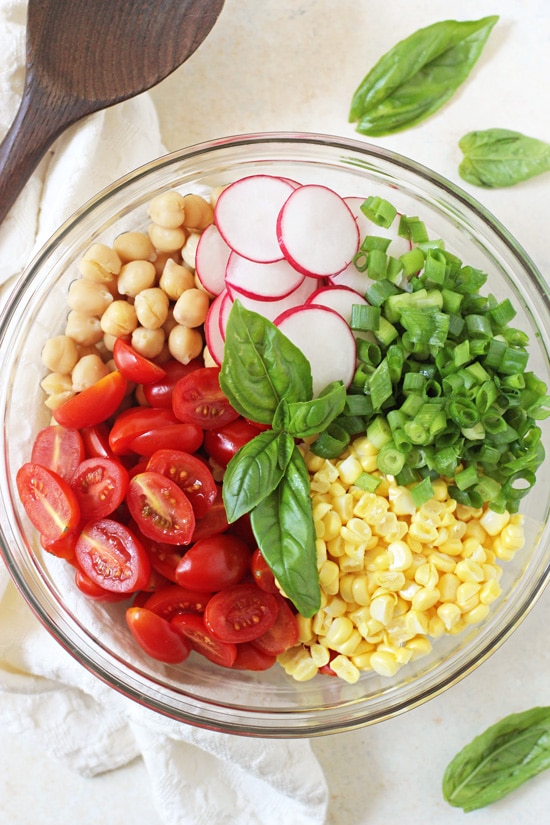 A glass bowl filled with chickpeas, tomatoes, corn, green onion and radishes.