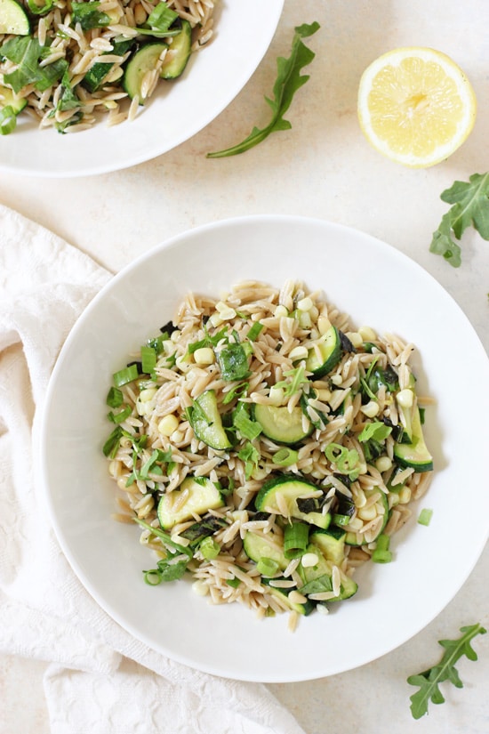 Two bowls of Herbed Zucchini and Corn Orzo Salad with a sliced lemon and white napkin.
