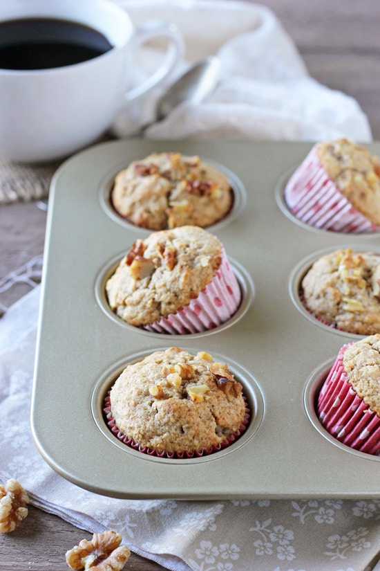 A side angle view of Vanilla Cardamom Walnut Muffins in a muffin tin with a cup of coffee in the background.