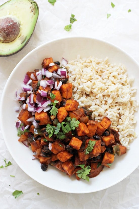 A white bowl filled with Sweet Potato & Black Bean Enchilada Stir-Fry and brown rice with an avocado to the side.