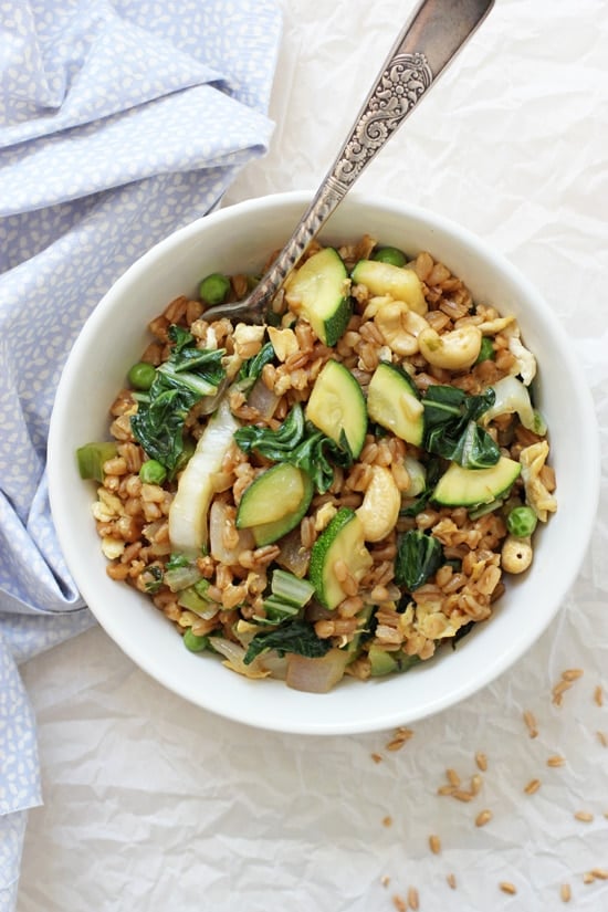 A white bowl of Green Veggie Farro Fried Rice with a fork and blue napkin to the side.