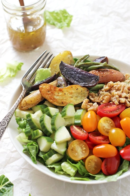 A side angle view of Greek Salad with Roasted Vegetables in a bowl with a fork and a jar of dressing in the background.