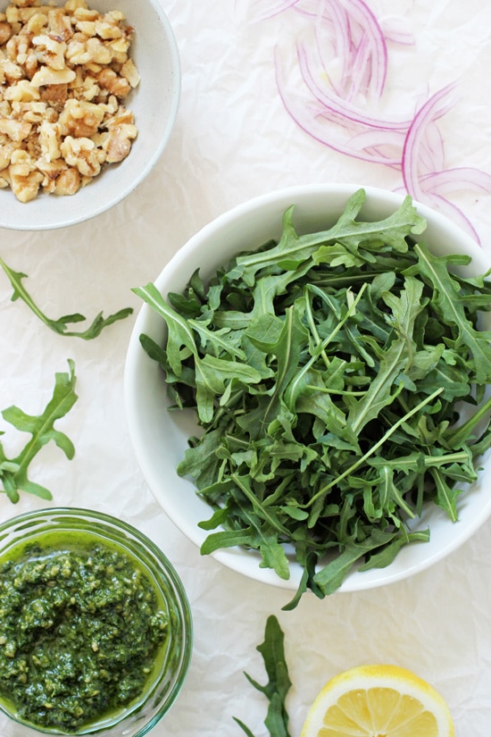 Small bowls of fresh arugula, chopped walnuts and pesto on crinkled white parchment paper.