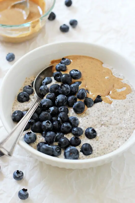 A white bowl with Blueberry Almond Butter Chia Pudding with a spoon and almond butter in the background.