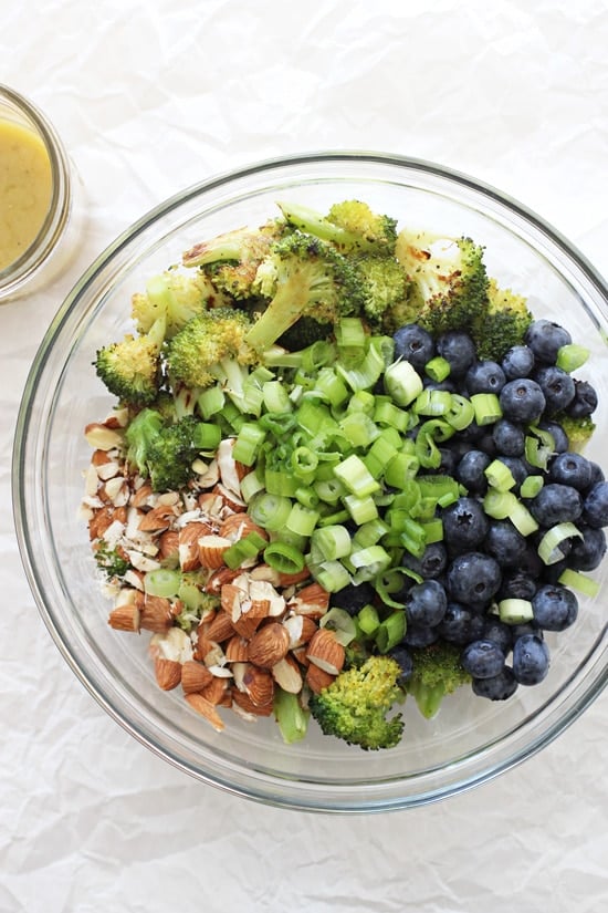 A glass bowl filled with roasted broccoli, blueberries, almonds and green onions.