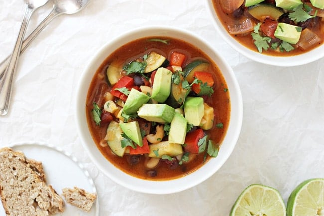 An overhead view of two bowls of Mexican Minestrone Soup with spoons, a lime and a slice of bread on the side.