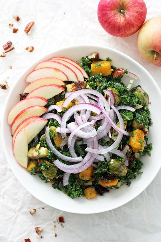 A large white bowl filled with Fall Harvest Kale Salad and two apples in the background.