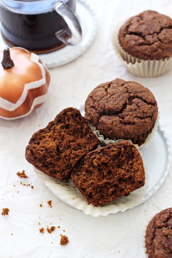 A side angle view of two Healthy Pumpkin Gingerbread Muffins with a cup of coffee and decorative pumpkin in the background.