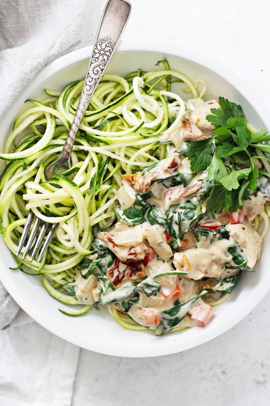 A large white bowl filled with Creamy Tuscan Garlic Chicken, zucchini noodles and a silver fork.