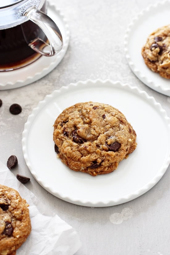 A Coconut Oil Oatmeal Cookie on a white plate with coffee and more cookies in the background.